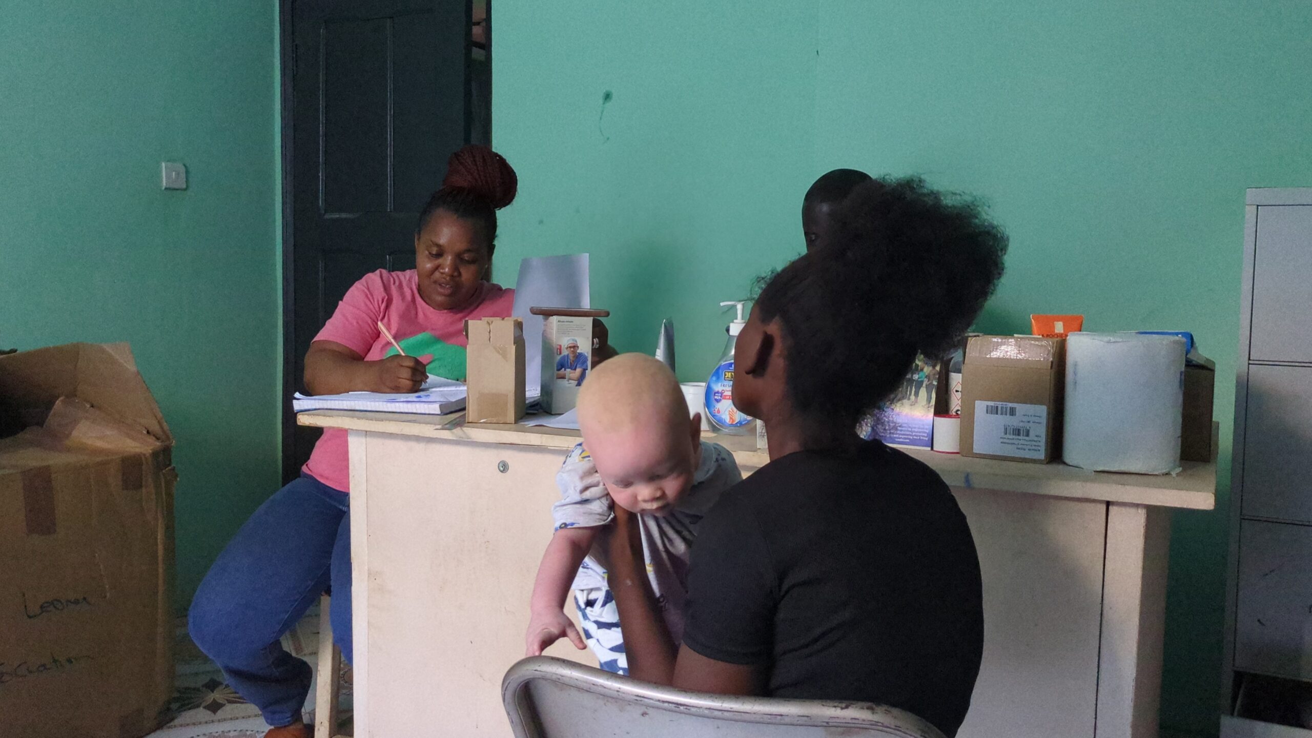 Nurse Jusu records information from a mother whose child has albinism at the SLAPWA clinic in Wellington, located in the east end of Freetown. This clinic serves as a gathering point for individuals with albinism, where they are screened monthly. Attendees are educated on measures to protect their skin from the sun, and they are also provided with sunscreen to shield their skin from the harmful effects of direct sunlight. Photo Credit: Kemo Cham, Engage Salone.